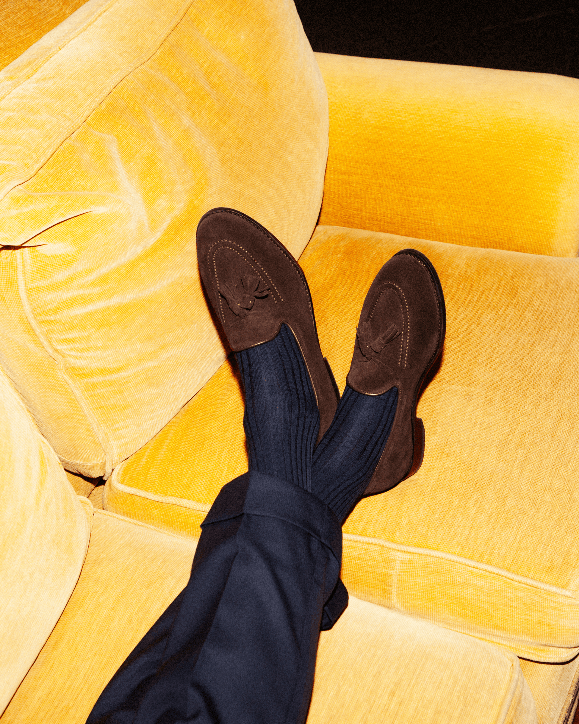 A man wearing Mölle II in dark brown suede relaxes on a yellow sofa, pairing the tassel loafers with navy dress socks and tailored navy slacks for a refined yet comfortable look.