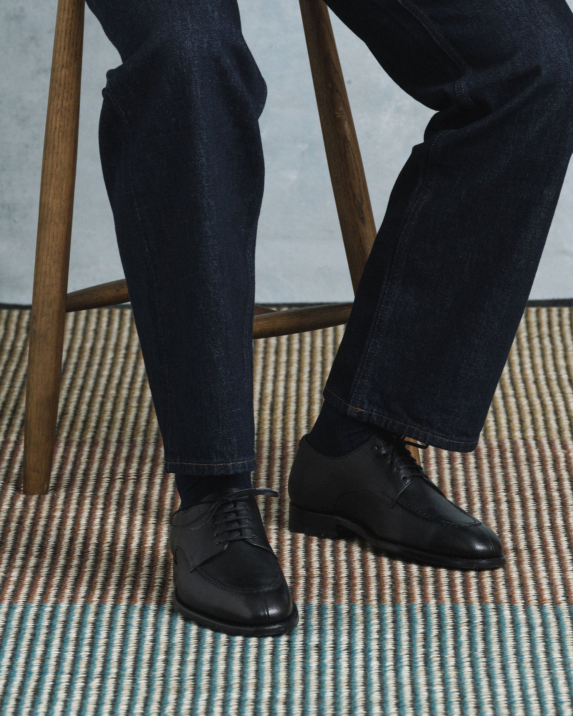 A man sits on a wooden stool wearing Norrmalm II in black hatch grain, styled with dark denim trousers, showcasing the textured split toe derby shoes in a relaxed, modern setting.