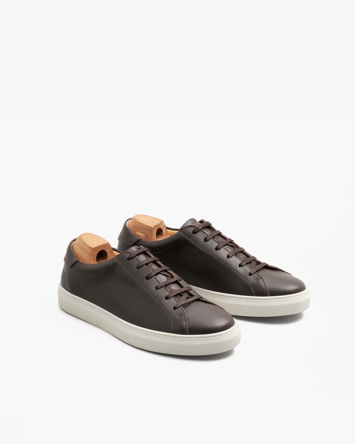 Oaxen in dark brown calf displayed at an angle, showcasing the smooth brown leather, clean stitching, and white rubber soles, each shoe fitted with a wooden shoe tree.