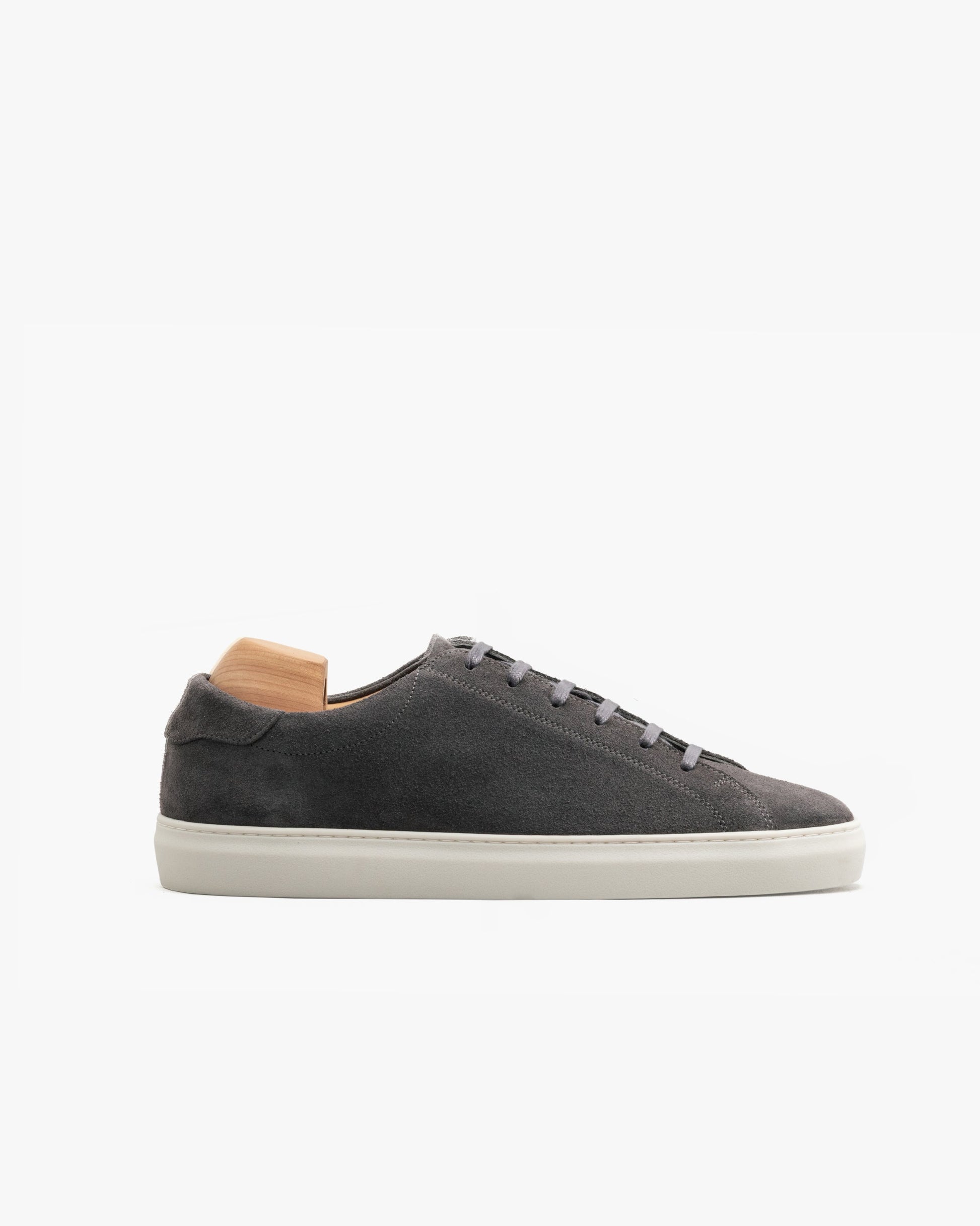 Low-top men’s sneaker in dark grey suede with a minimalist silhouette, tonal stitching, flat laces, and an off-white sole, shown in profile with a wooden shoe tree inside.