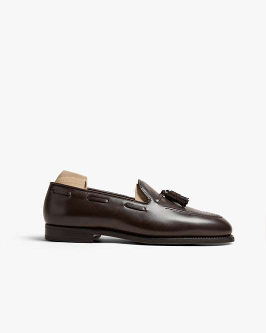 A single Mölle II loafer in dark brown calf leather is displayed in profile, highlighting its polished finish, apron front, and refined tassel detail, set on a classic leather sole with a wooden shoe tree inside.