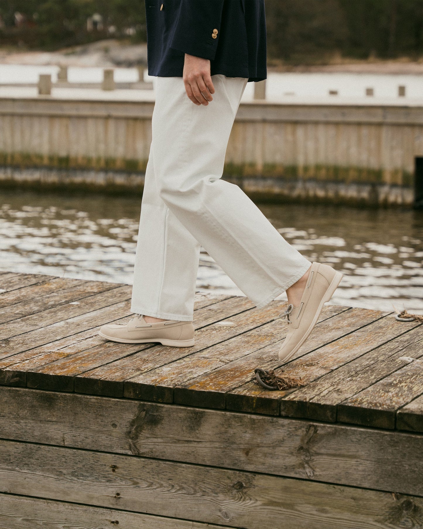 A man walks along a wooden dock wearing Sandhamn in beige nubuck, styled with white slacks and a navy blazer, highlighting the boat shoe’s relaxed fit and refined, minimalist design in a coastal setting.
