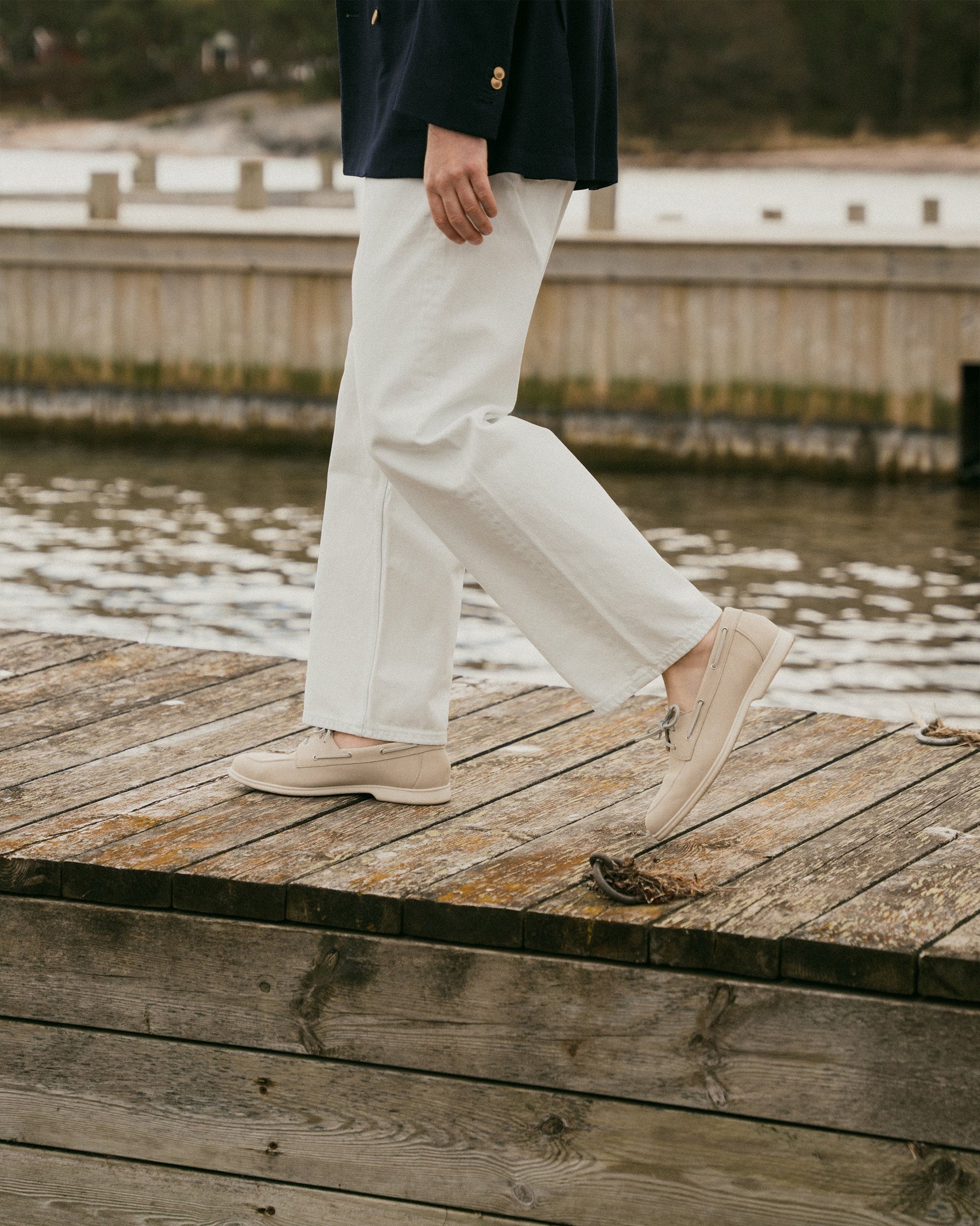 A man walks along a wooden dock wearing Sandhamn in beige nubuck, styled with white slacks and a navy blazer, highlighting the boat shoe’s relaxed fit and refined, minimalist design in a coastal setting.