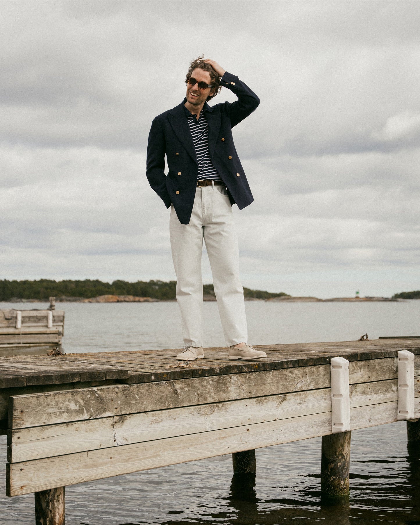 A man stands on a wooden dock by the water wearing Sandhamn in beige nubuck, styled with white slacks, a navy blazer, and a striped shirt, highlighting the boat shoe’s relaxed fit and understated Scandinavian-inspired design for casual summer occasions.