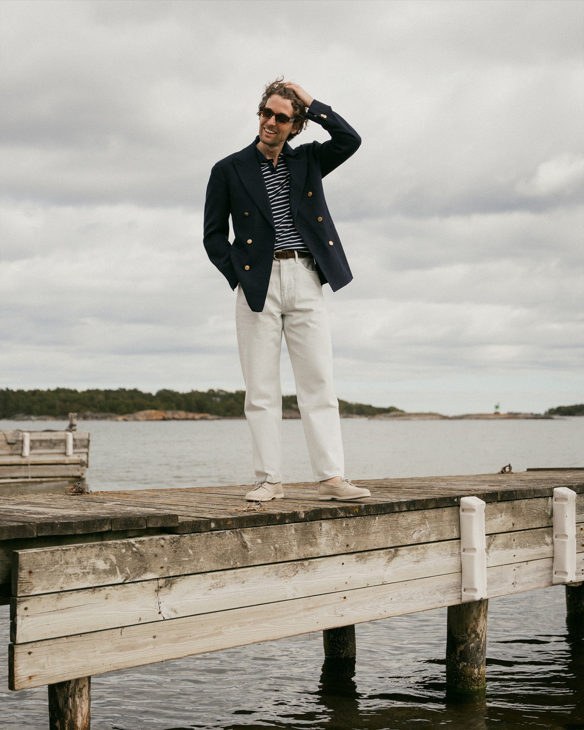 A man stands on a wooden dock by the water wearing Sandhamn in beige nubuck, styled with white slacks, a navy blazer, and a striped shirt, highlighting the boat shoe’s relaxed fit and understated Scandinavian-inspired design for casual summer occasions.