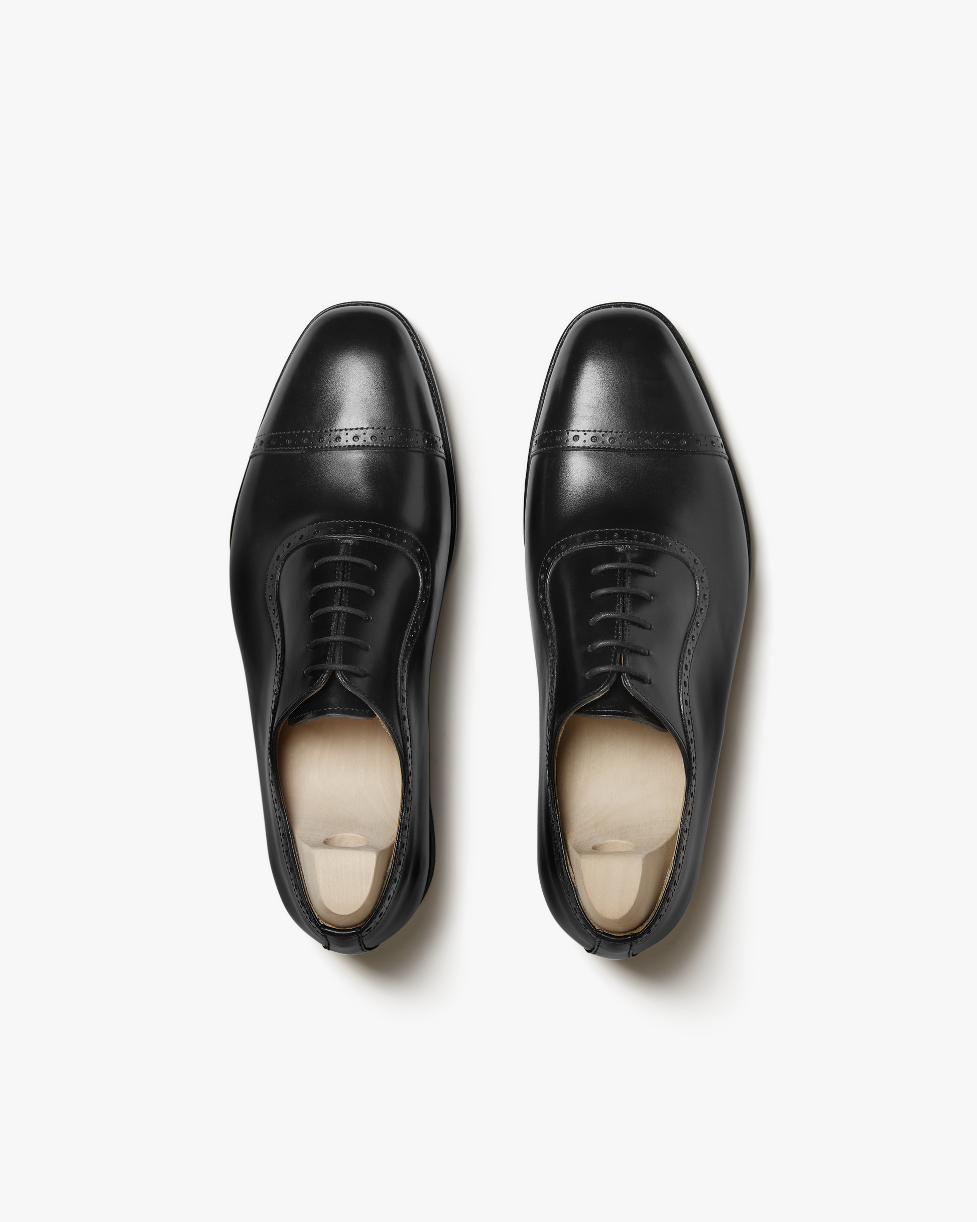 Pair of Skytteholm II in black calf shown from above, highlighting the closed lacing, gently chiseled toe, and subtle brogue perforations on the cap, with wooden shoe trees inside each Oxford.