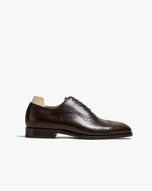 Dark brown oxford shoe in polished calf leather with cap toe and subtle brogue detailing, shown in profile against a white background. The closed lacing and gently chiseled toe highlight its refined silhouette, ideal for pairing with suits or formal slacks.