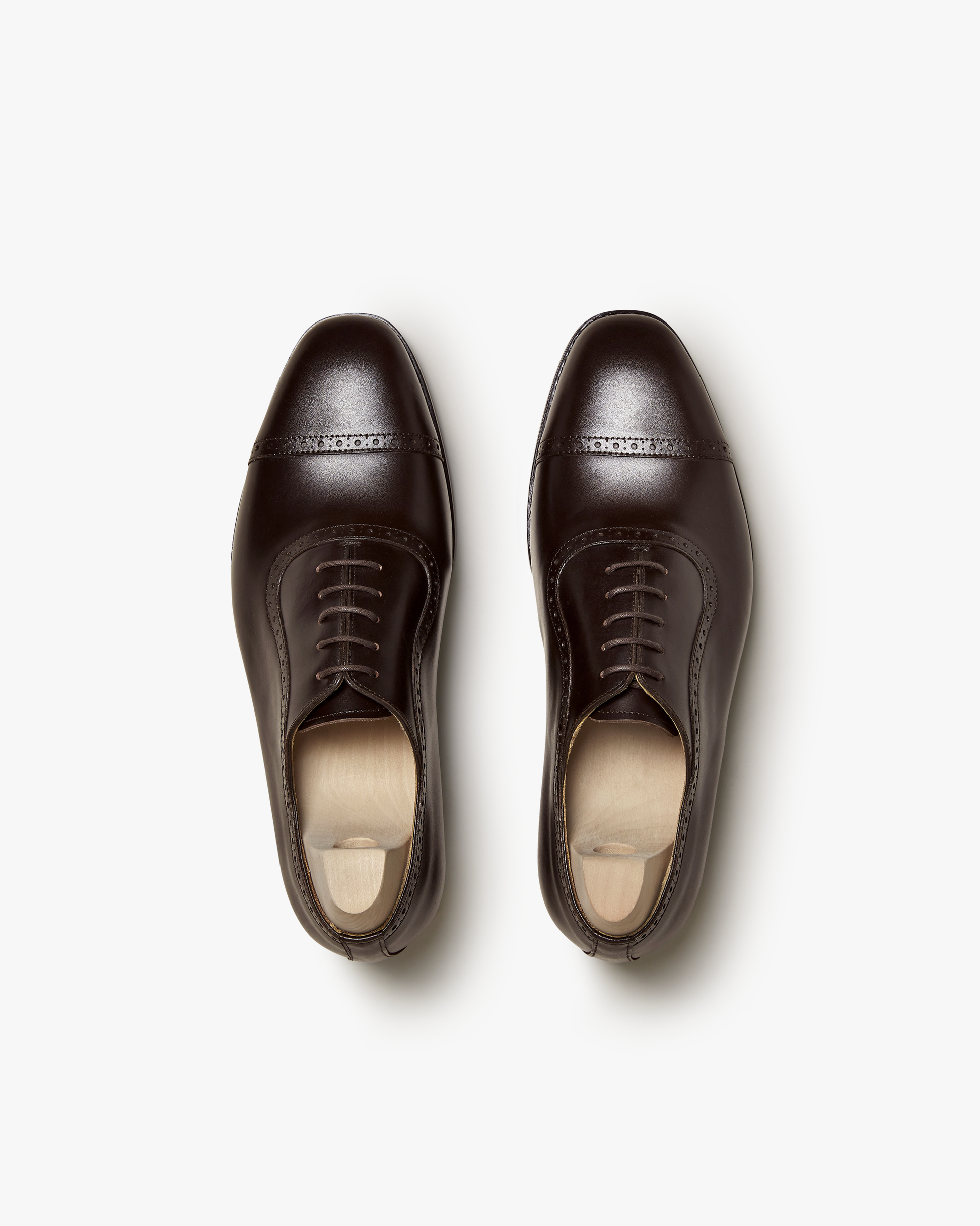 Pair of Skytteholm II oxford shoes in dark brown calf, shown from above to highlight the cap toe, subtle brogue detailing, and elegant closed lacing, with wooden shoe trees inside each shoe.