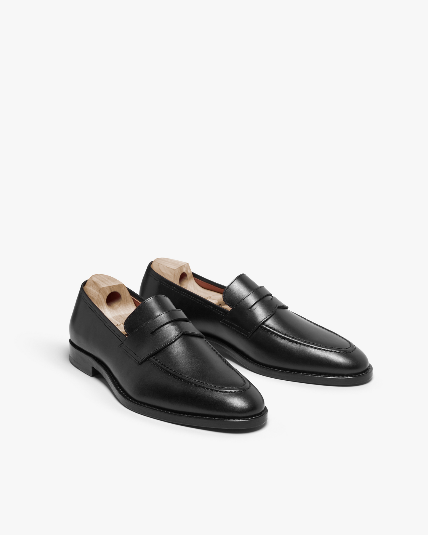 Smögen loafers in black calf leather feature a classic penny strap, gently rounded toe, and smooth finish, shown at an angle with wooden shoe trees inside each shoe.