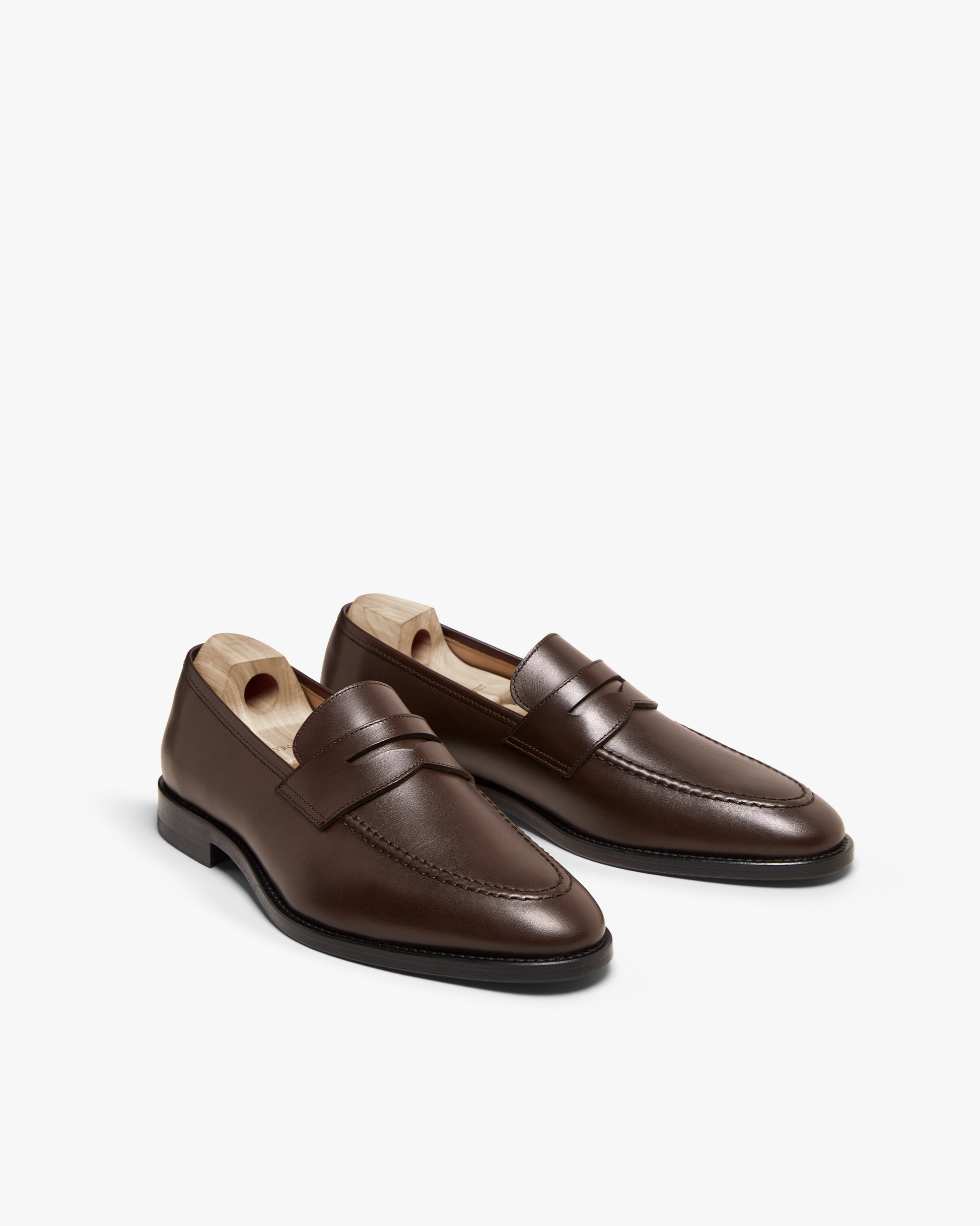 Smögen loafers in dark brown calf leather displayed at an angle, featuring a classic penny strap, gently rounded toe, and smooth finish, with wooden shoe trees inside to maintain their shape.