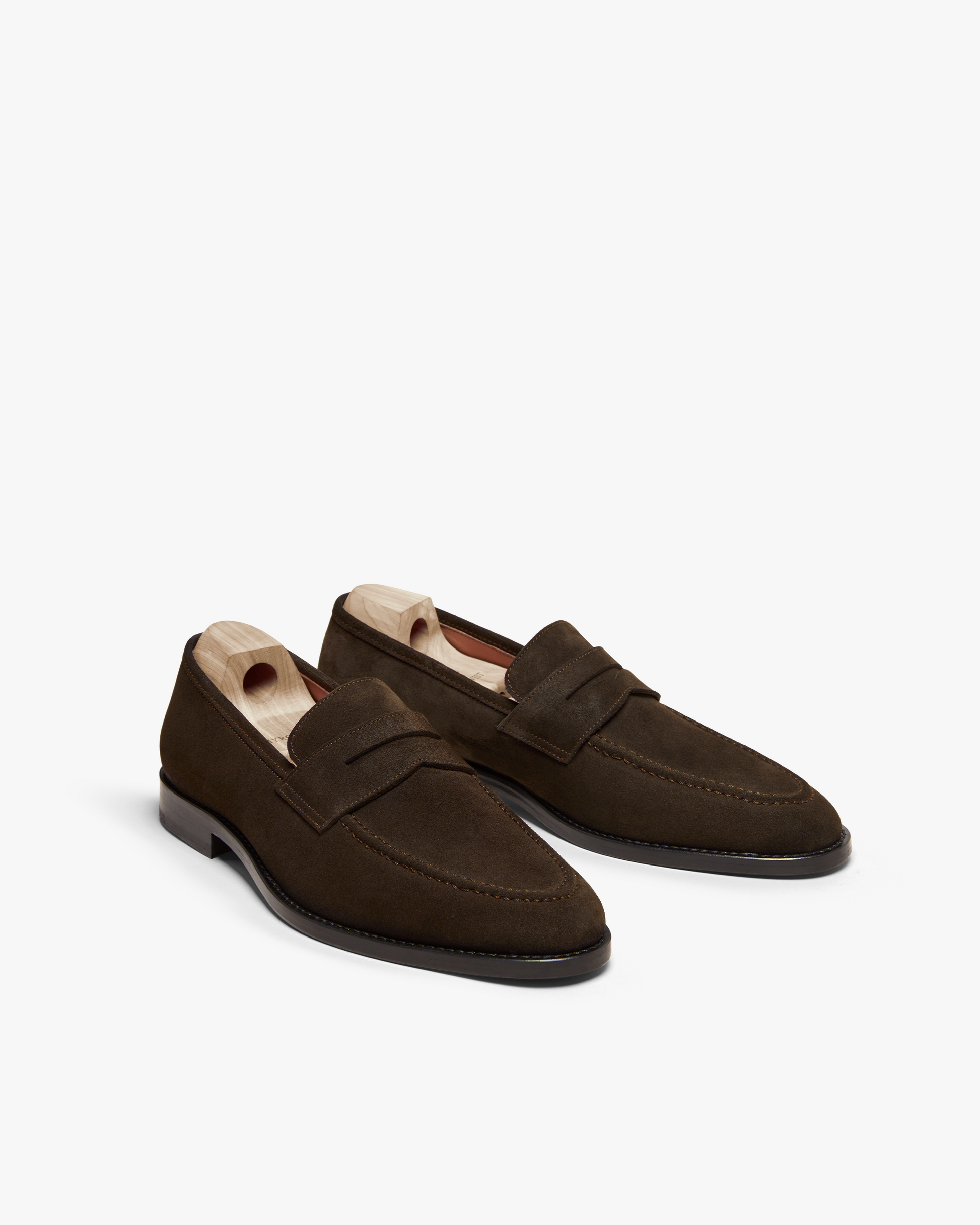 Smögen loafers in dark brown suede with a classic penny strap and softly rounded toe, shown with wooden shoe trees to preserve their shape and highlight the rich suede texture and refined stitching.