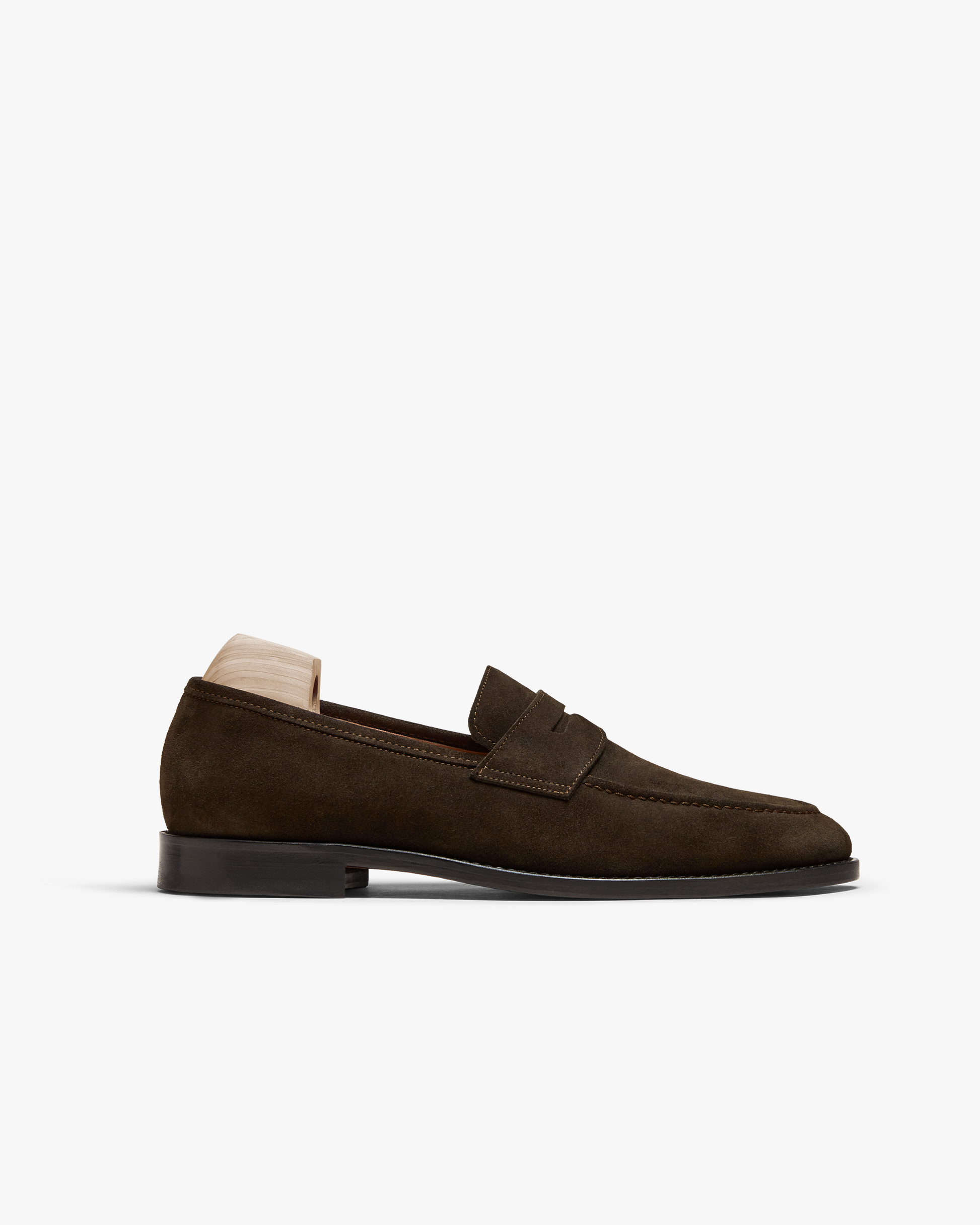 Smögen loafer in dark brown suede features a softly rounded toe, classic penny strap, and subtle stitching, shown with a wooden shoe tree to maintain its elegant shape. The rich suede finish and low stacked heel highlight its versatile style, ideal for pairing with tailored slacks or relaxed suiting.