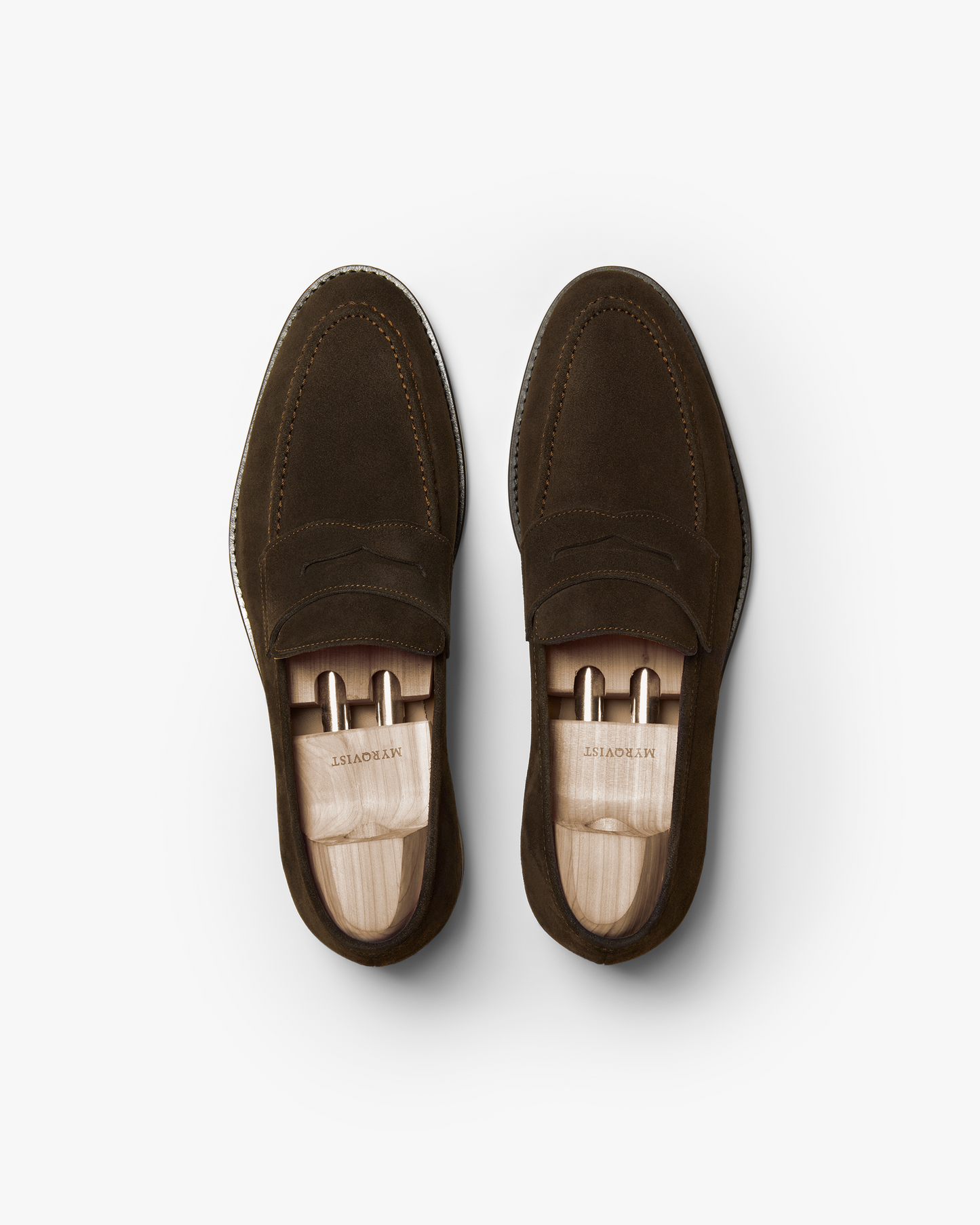 Pair of Smögen loafers in dark brown suede shown from above, highlighting the soft suede texture, classic penny strap design, and wooden shoe trees that help maintain their shape. The stitching and rounded toe offer a refined look suitable for both business and smart casual attire.