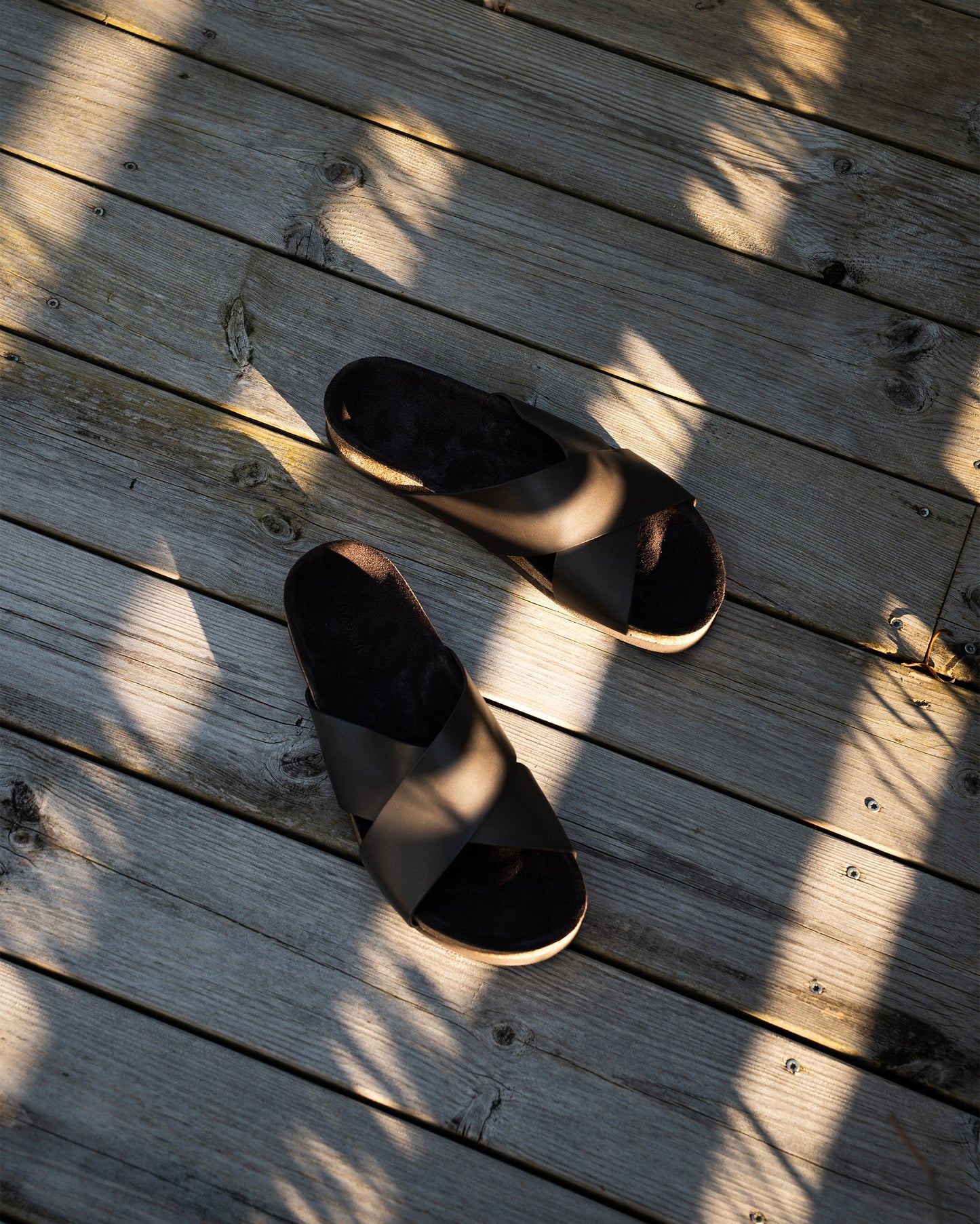 Solvik in dark brown calf with wide, criss-crossed leather straps and a cork sole is shown resting on sunlit wooden decking, highlighting its relaxed summer style and refined, minimal design.