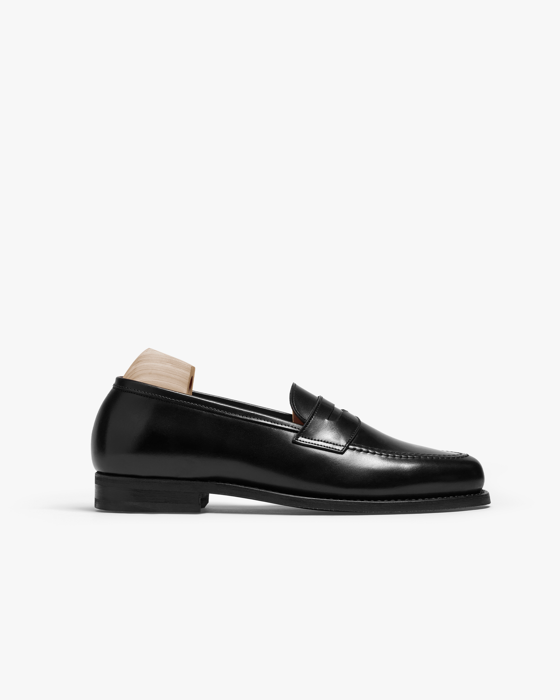 Stenhammar II in black calf features a polished penny loafer silhouette with an apron front, rounded toe, and subtle stitching, shown with a wooden shoe tree to maintain its refined shape.