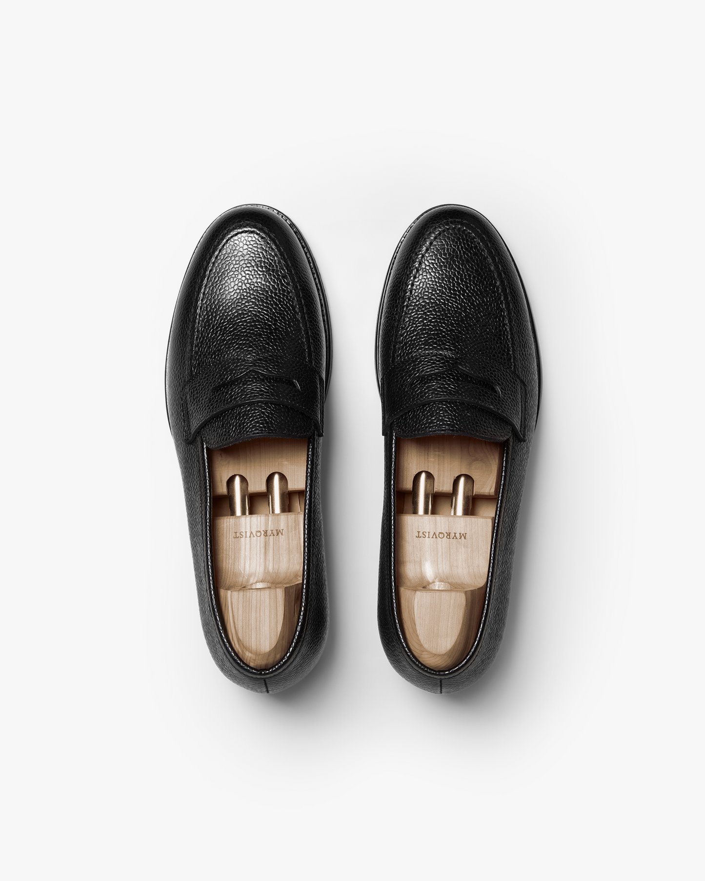 Stenhammar II in black country calf shown from above, featuring textured grain leather, penny strap detailing, and wooden shoe trees inside each loafer.