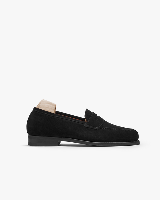 Stenhammar II in black suede shown in profile, featuring a classic penny loafer design with an apron front, rounded toe, and a full rubber sole, presented with a wooden shoe tree inside.