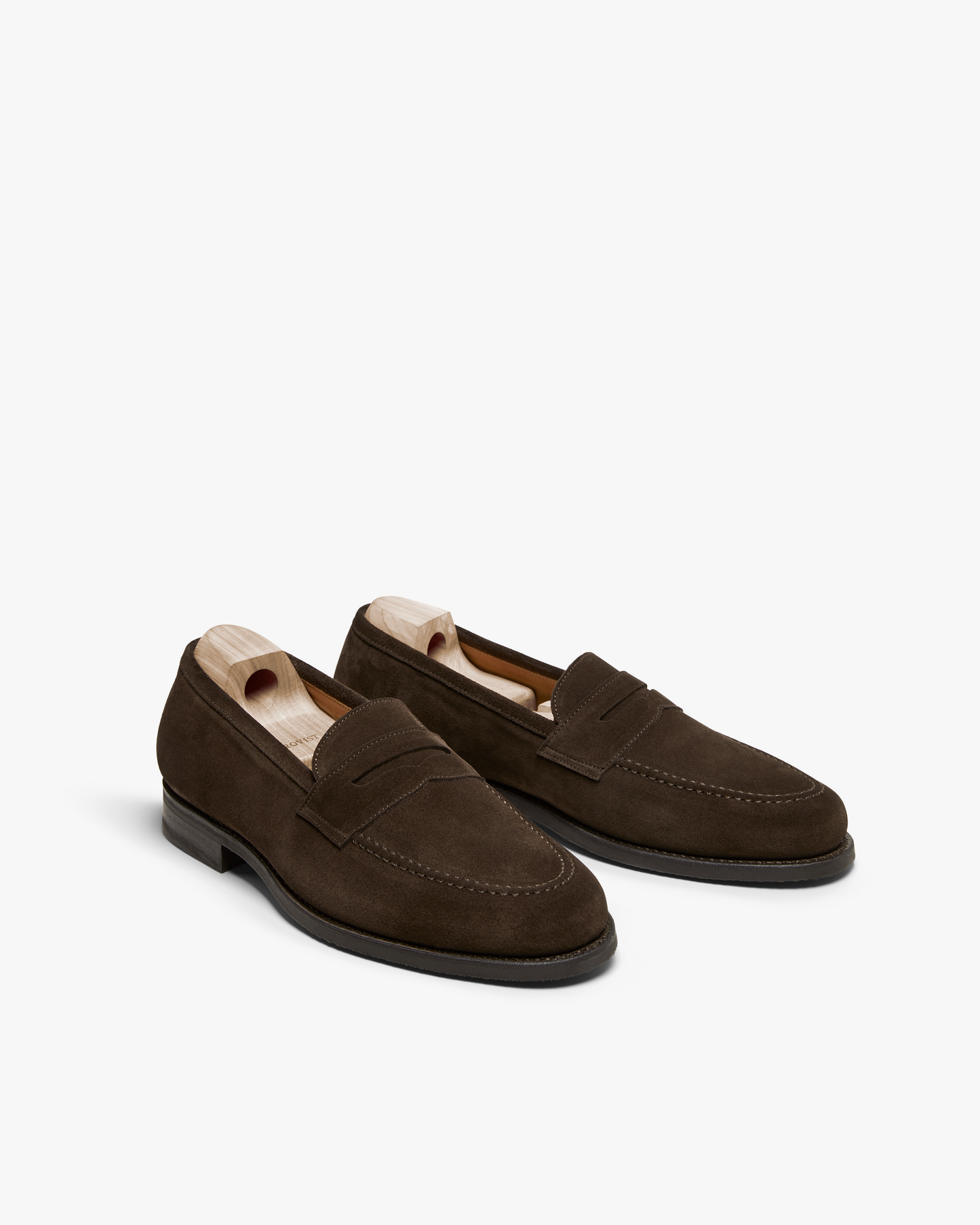 Stenhammar II in dark brown suede displayed at an angle, featuring a classic penny loafer design with apron stitching, rich suede texture, and wooden shoe trees inside each shoe.