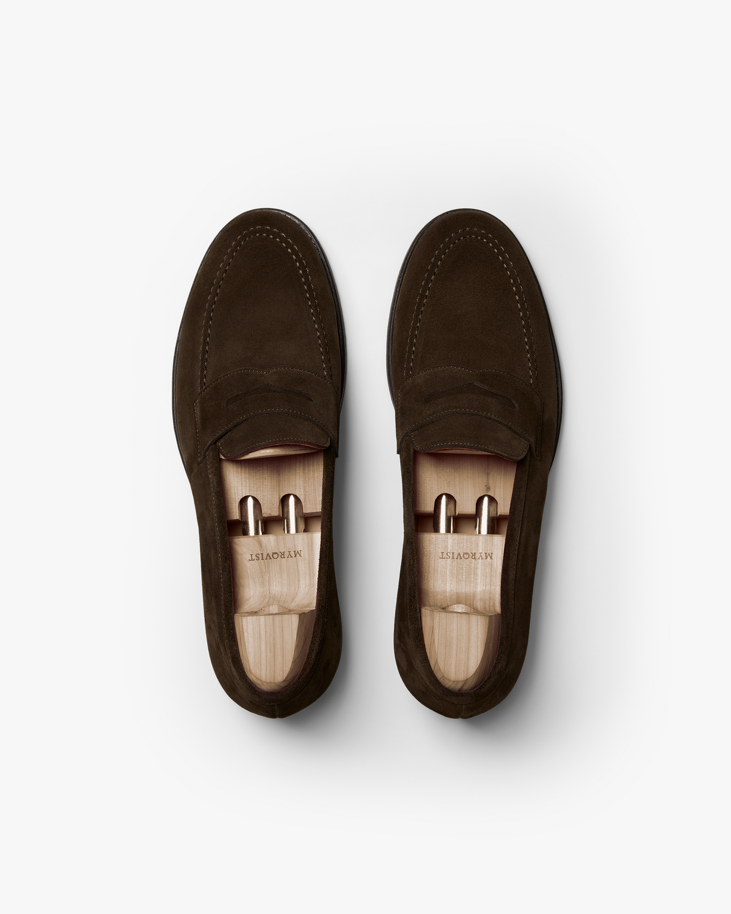 A pair of Stenhammar II in dark brown suede shown from above, featuring a softly rounded toe, apron stitching, and wooden shoe trees inside each loafer.