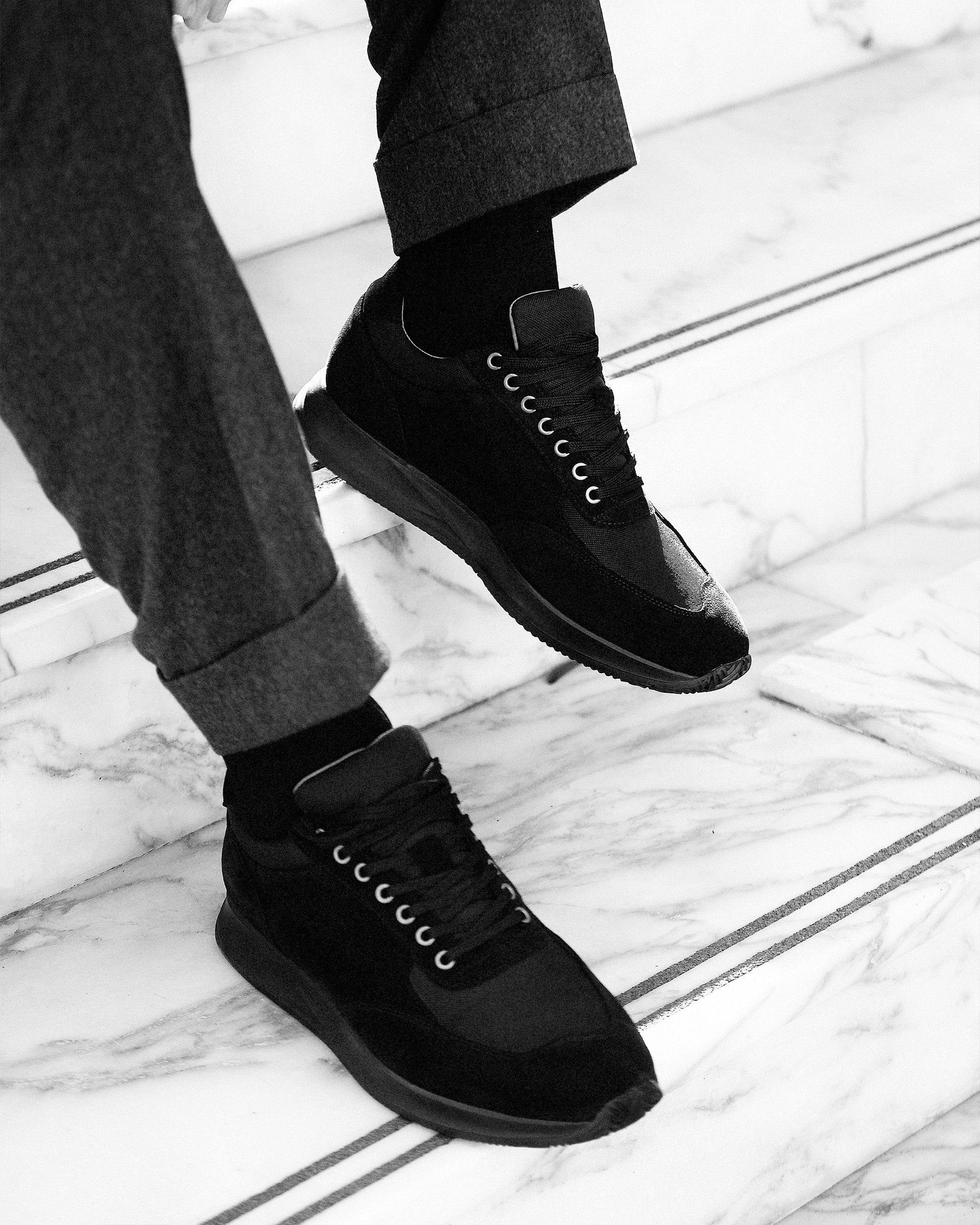 A man wearing Stensund in black suede, styled with tailored slacks, steps confidently on marble stairs, highlighting the trainer’s sleek silhouette, tonal mesh panels, and versatile design suited for both business casual and relaxed settings.
