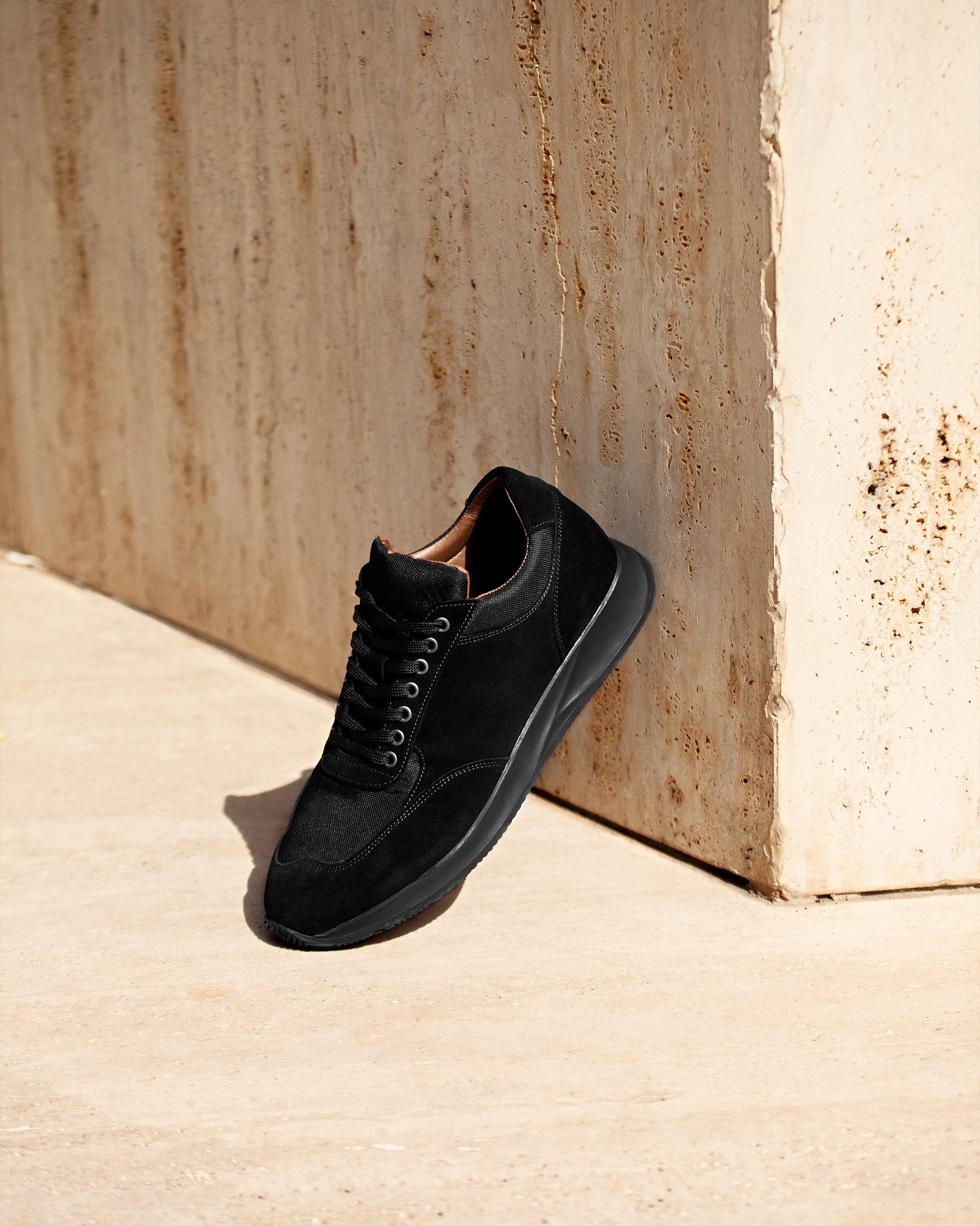 Stensund in black suede displayed outdoors against a textured stone wall, showcasing its clean silhouette, tonal mesh panels, and refined stitching for a versatile and modern trainer.