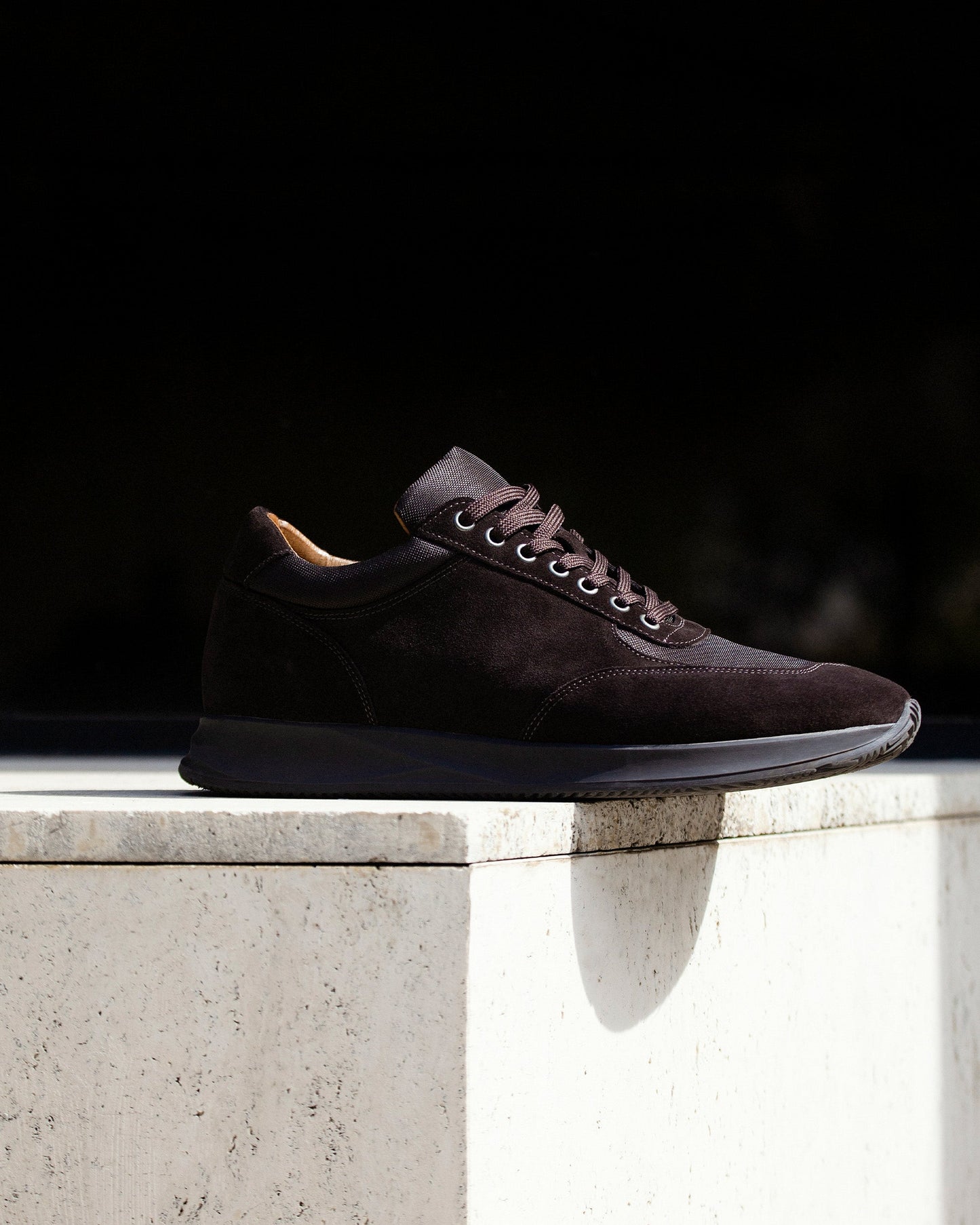 Stensund in dark brown suede displayed on a light stone surface, showcasing its smooth suede upper, tonal mesh accents, and sculpted rubber sole in natural sunlight.