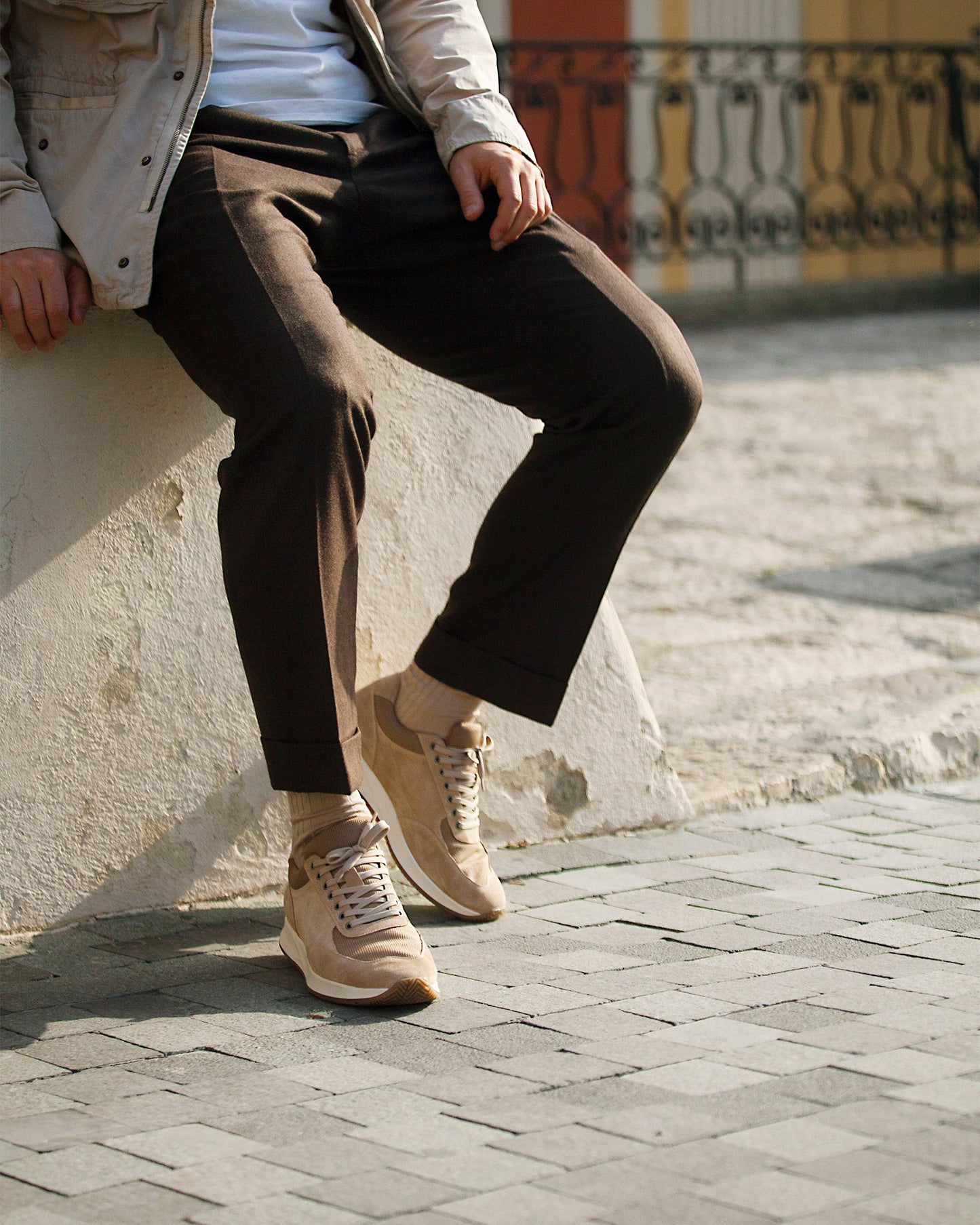 A man sits outdoors wearing Stensund in sand suede, styled with brown slacks and a light jacket, highlighting the trainer's soft beige suede, tonal mesh panels, and versatile design suited for both business casual and relaxed settings.
