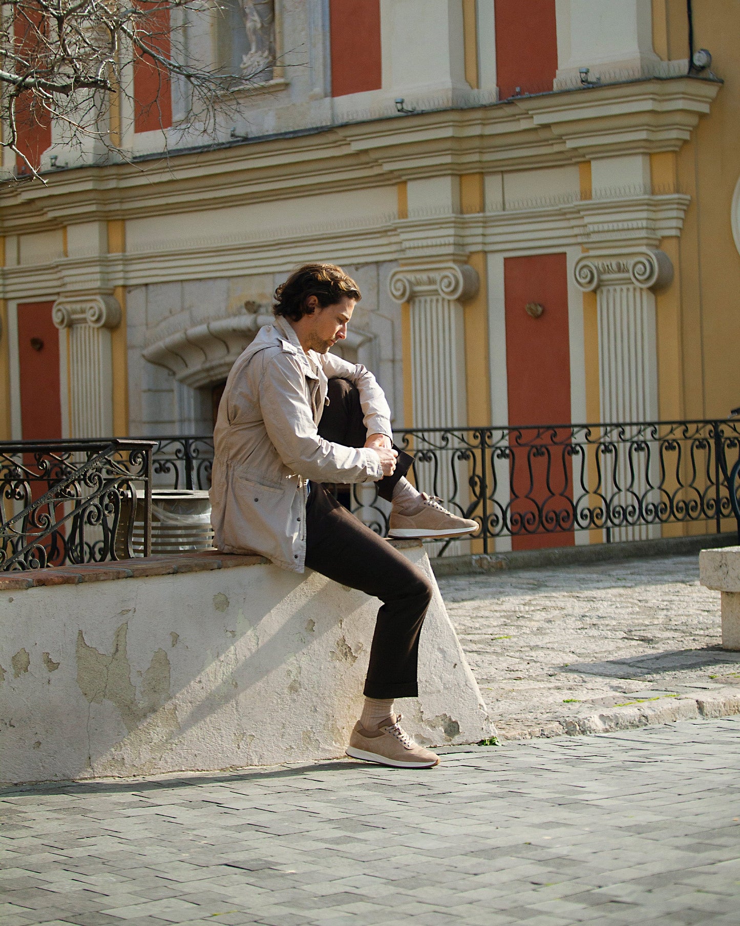 A man sits on a stone ledge in front of a historic building, wearing Stensund in sand suede with brown slacks and a light jacket, highlighting the trainer's soft beige suede and relaxed, versatile style.