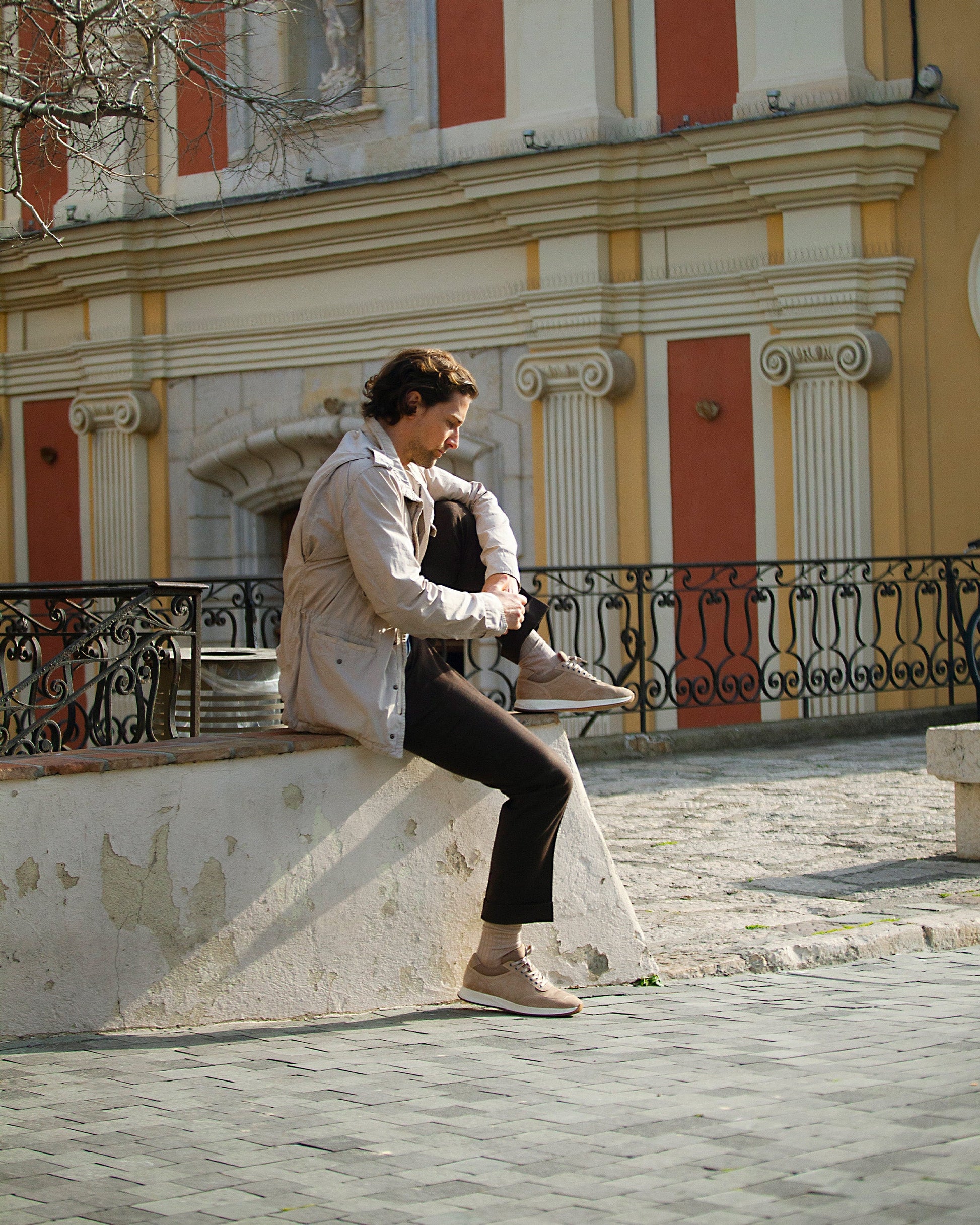 A man sits on a stone ledge in front of a historic building, wearing Stensund in sand suede with brown slacks and a light jacket, highlighting the trainer's soft beige suede and relaxed, versatile style.