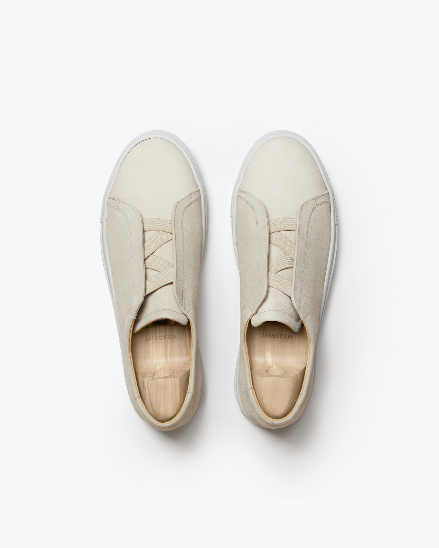 Top view of Alnö II in beige nubuck showing the seamless slip-on design with elastic crossover detail, softly textured uppers, and wooden shoe trees inside each sneaker.