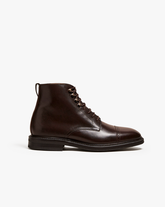 A dark brown calf leather men's boot with a rounded toe, subtle broguing on the cap toe, metal lace hooks, and a pull tab at the heel, shown in a side profile on a white background. The boot features a smooth finish and classic silhouette, highlighting its refined and versatile design.
