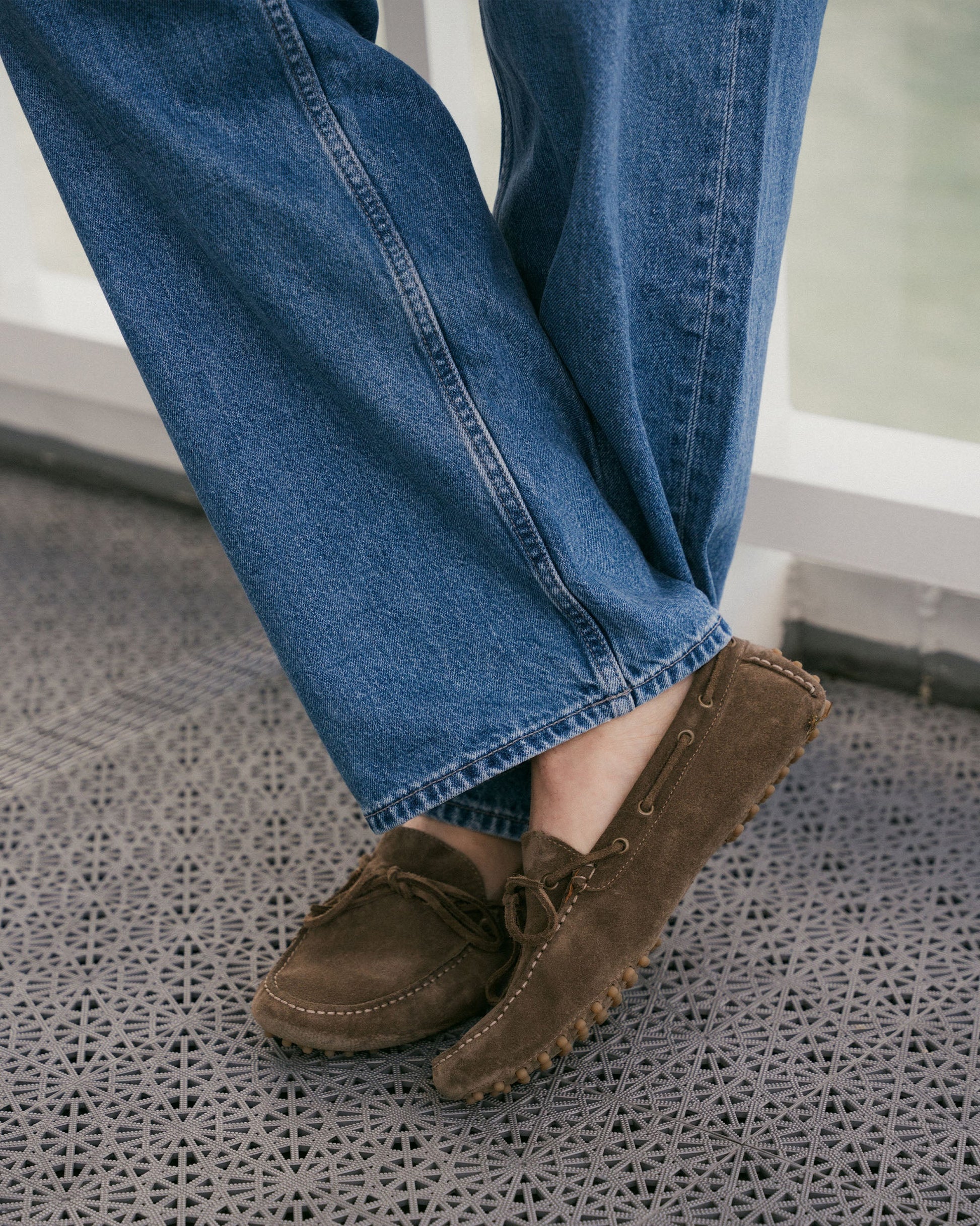 A pair of Tofta loafers in taupe suede, featuring a rubber driving sole and subtle bow detail, worn with relaxed blue denim trousers in a casual setting.