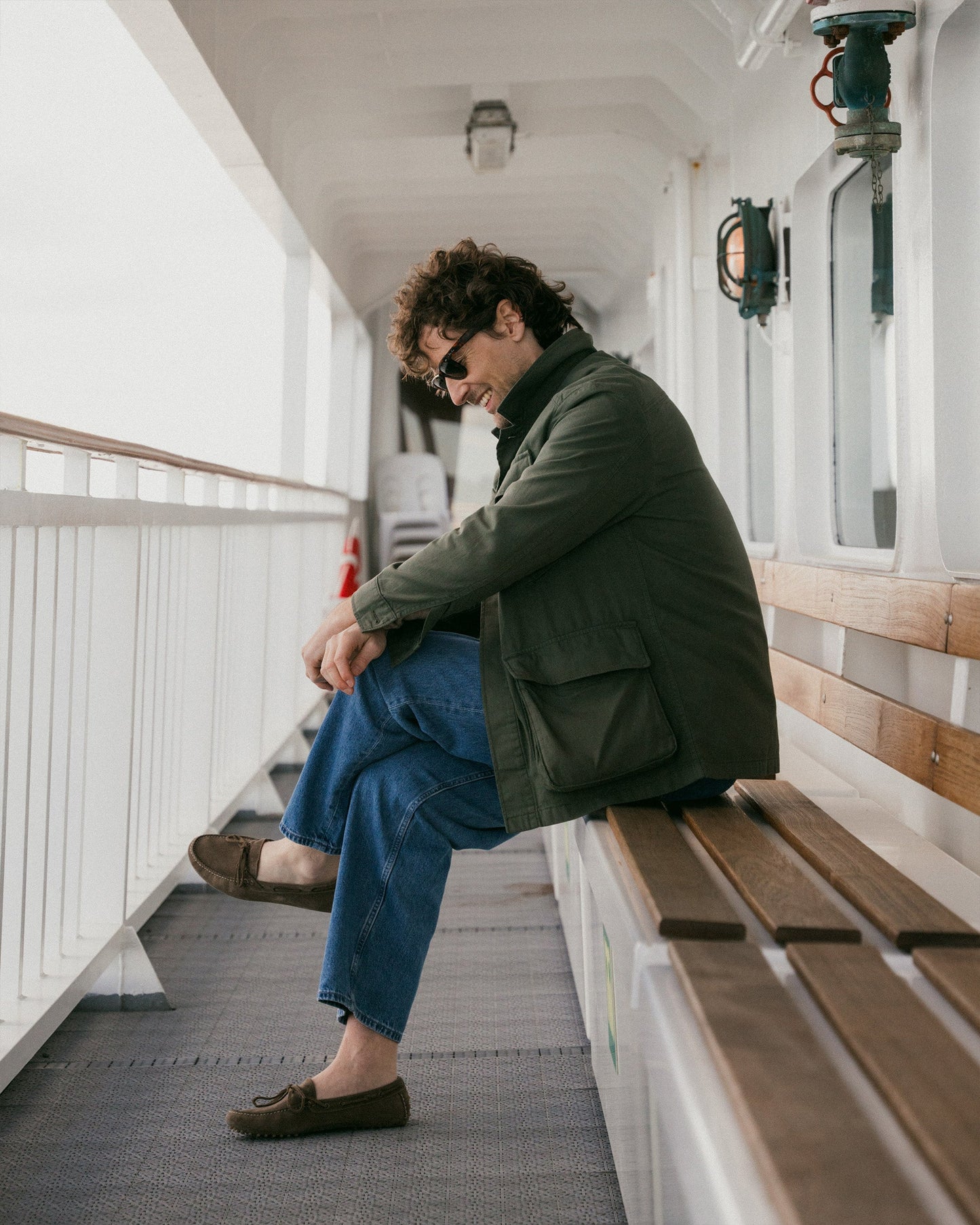A man sits on a wooden bench aboard a ferry, wearing Tofta loafers in taupe suede paired with relaxed blue denim trousers and an olive green jacket, highlighting the loafers’ casual versatility and understated style.