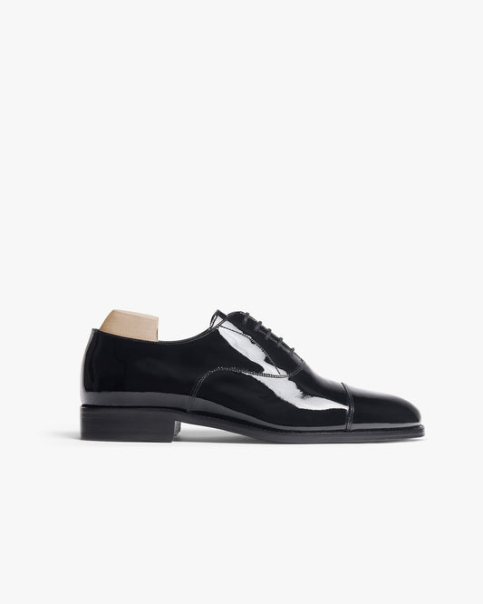 Black patent leather oxford shoe with a softly rounded cap toe, mirror-like finish, and subtle stitching, displayed in profile with a wooden shoe tree inside.