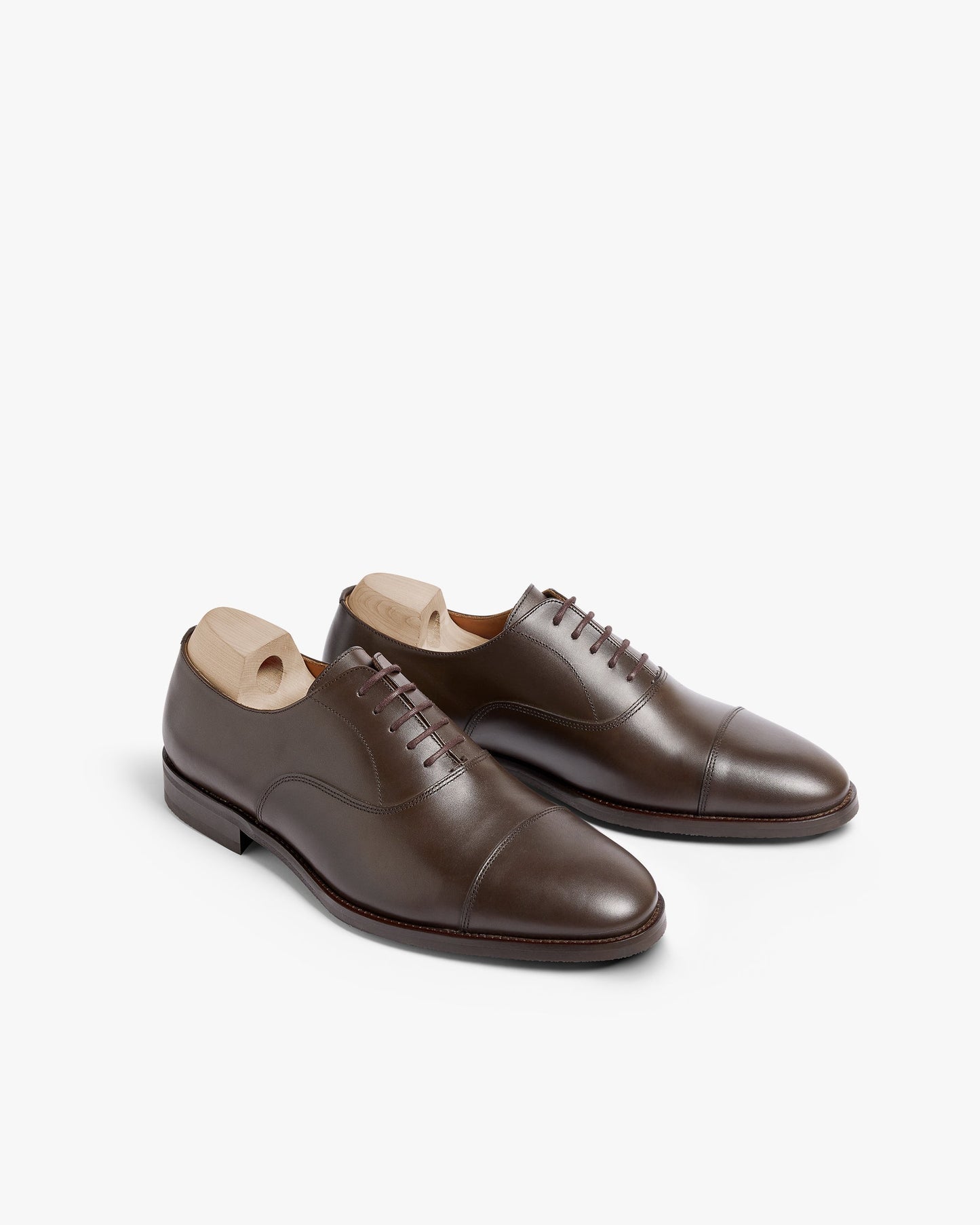 Vinterviken II in dark brown calf, a pair of classic oxford shoes featuring gently chiseled cap toes, clean stitching, and wooden shoe trees, displayed at an angle on a white background.