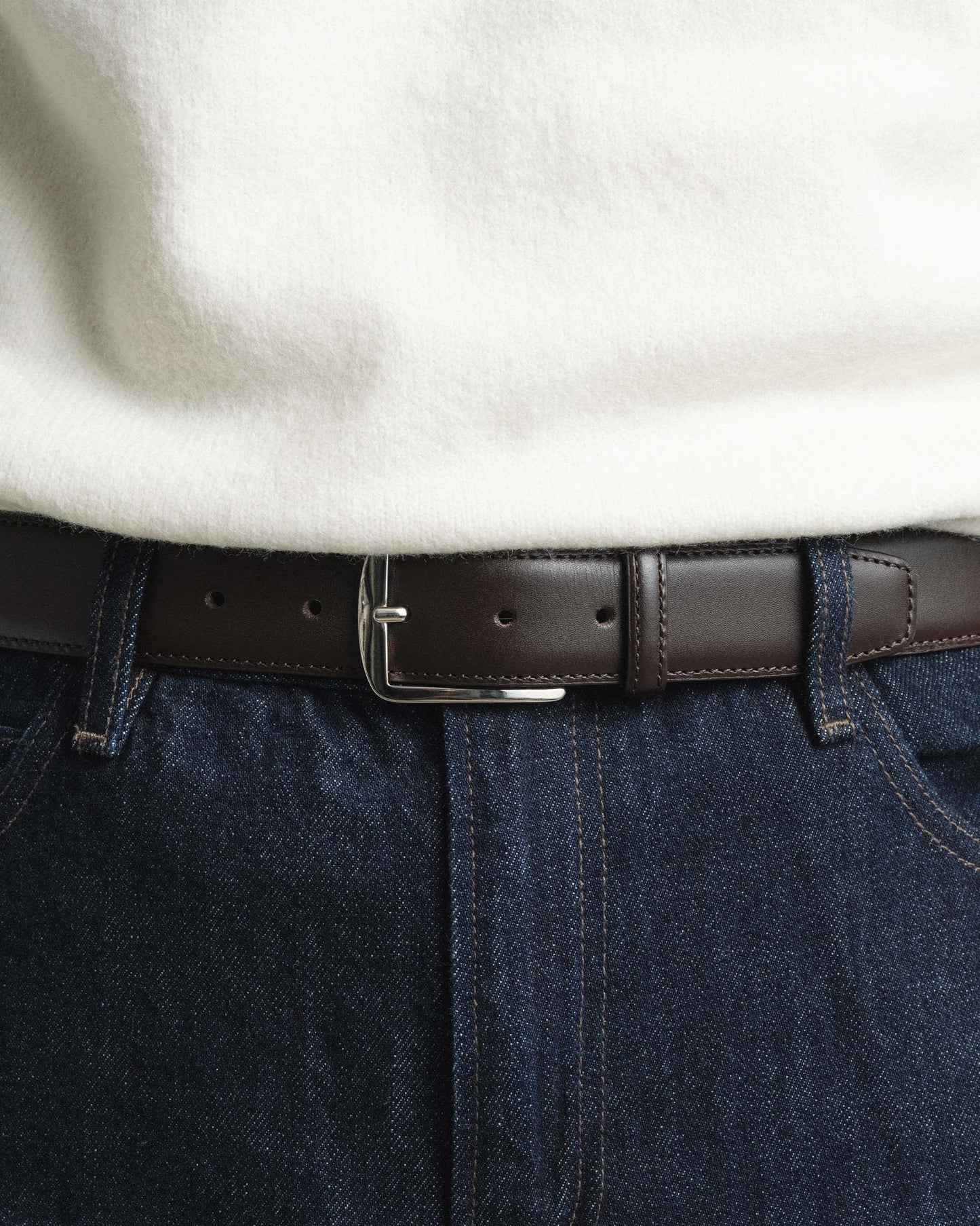 Men’s dark brown leather belt with a silver square buckle, styled with dark denim jeans and a cream sweater for a refined, versatile look.