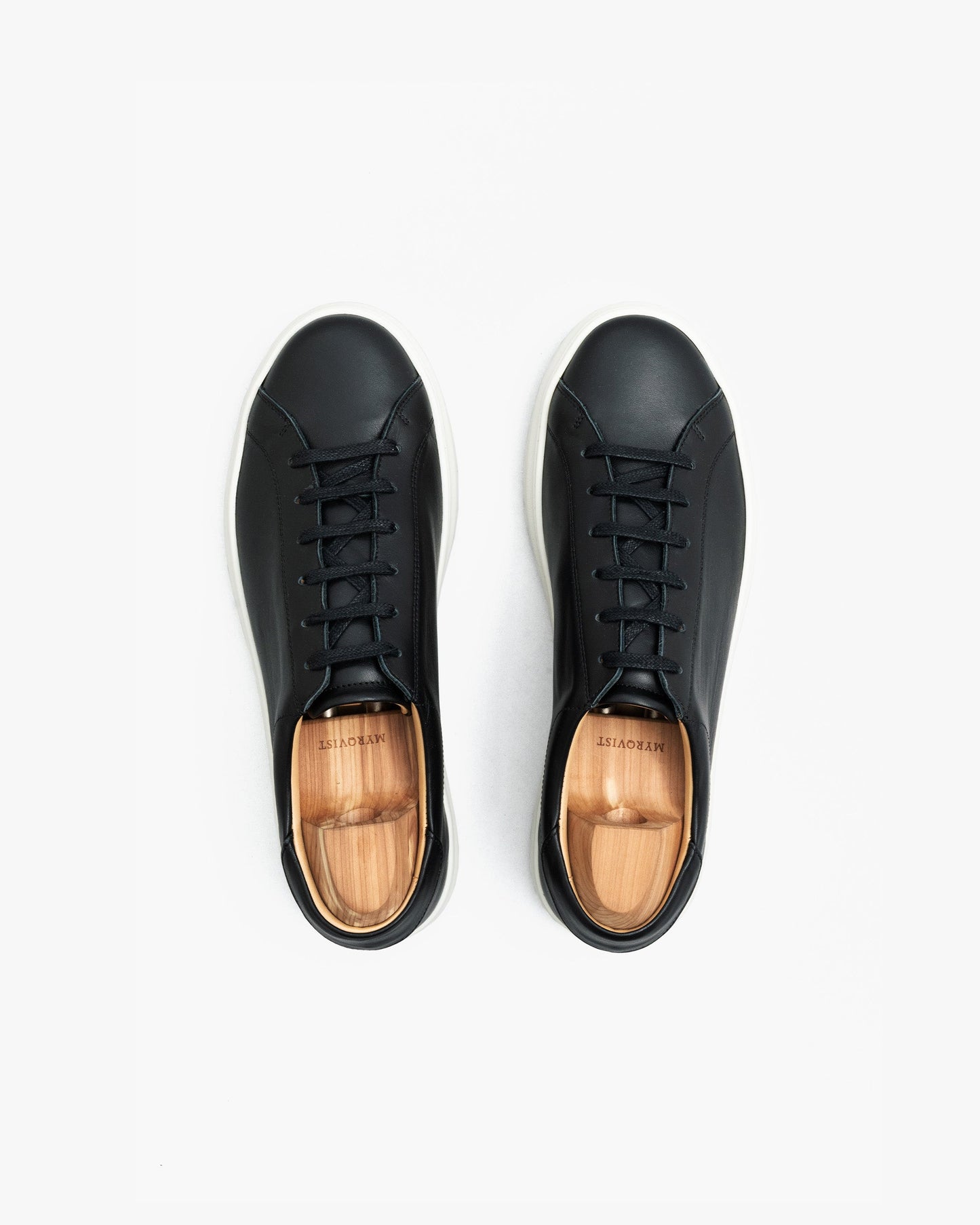 Pair of Oaxen sneakers in black calf leather shown from above, featuring clean stitching, black laces, white soles, and wooden shoe trees inside each shoe.