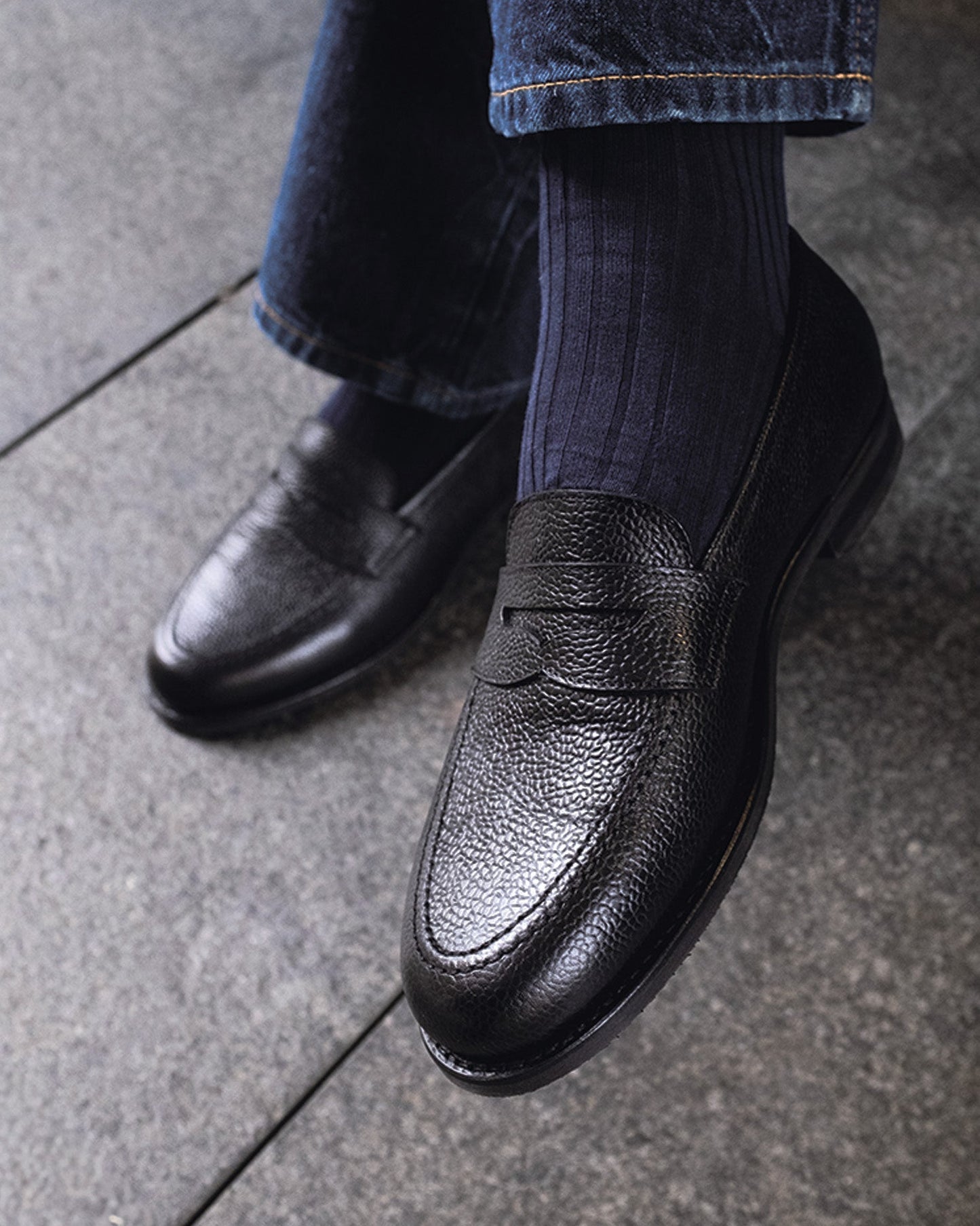 Stenhammar II in black country calf worn with dark socks and denim, showcasing the textured grain leather, penny strap, and apron toe design in a casual setting.