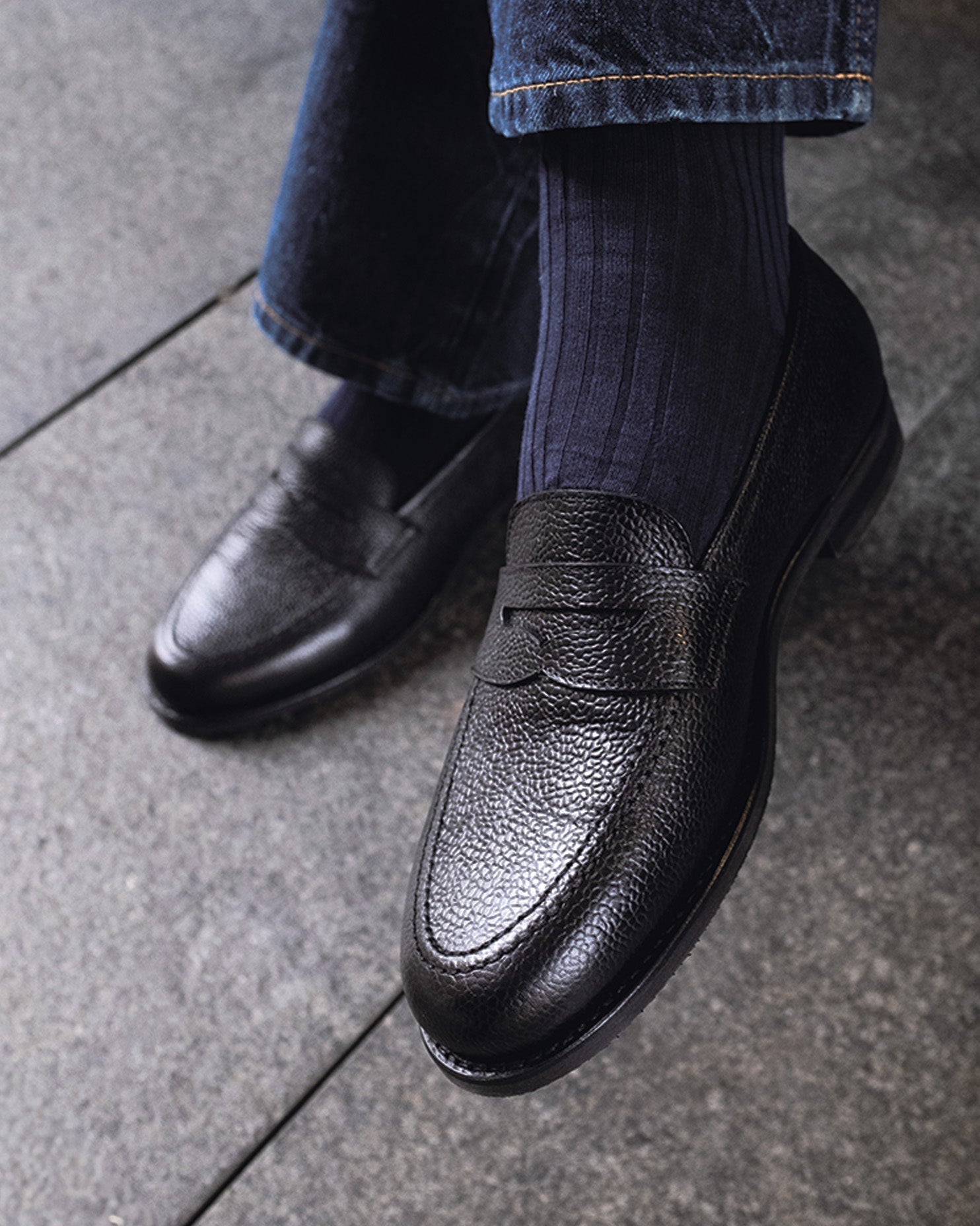 Stenhammar II in black country calf worn with dark socks and denim, showcasing the textured grain leather, penny strap, and apron toe design in a casual setting.