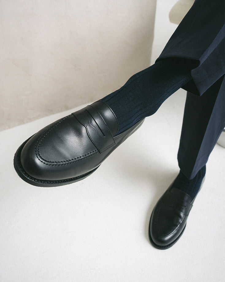 A man wears Stenhammar II in black calf, styled with dark socks and tailored navy slacks, showcasing the loafer’s polished apron front and timeless penny strap design.