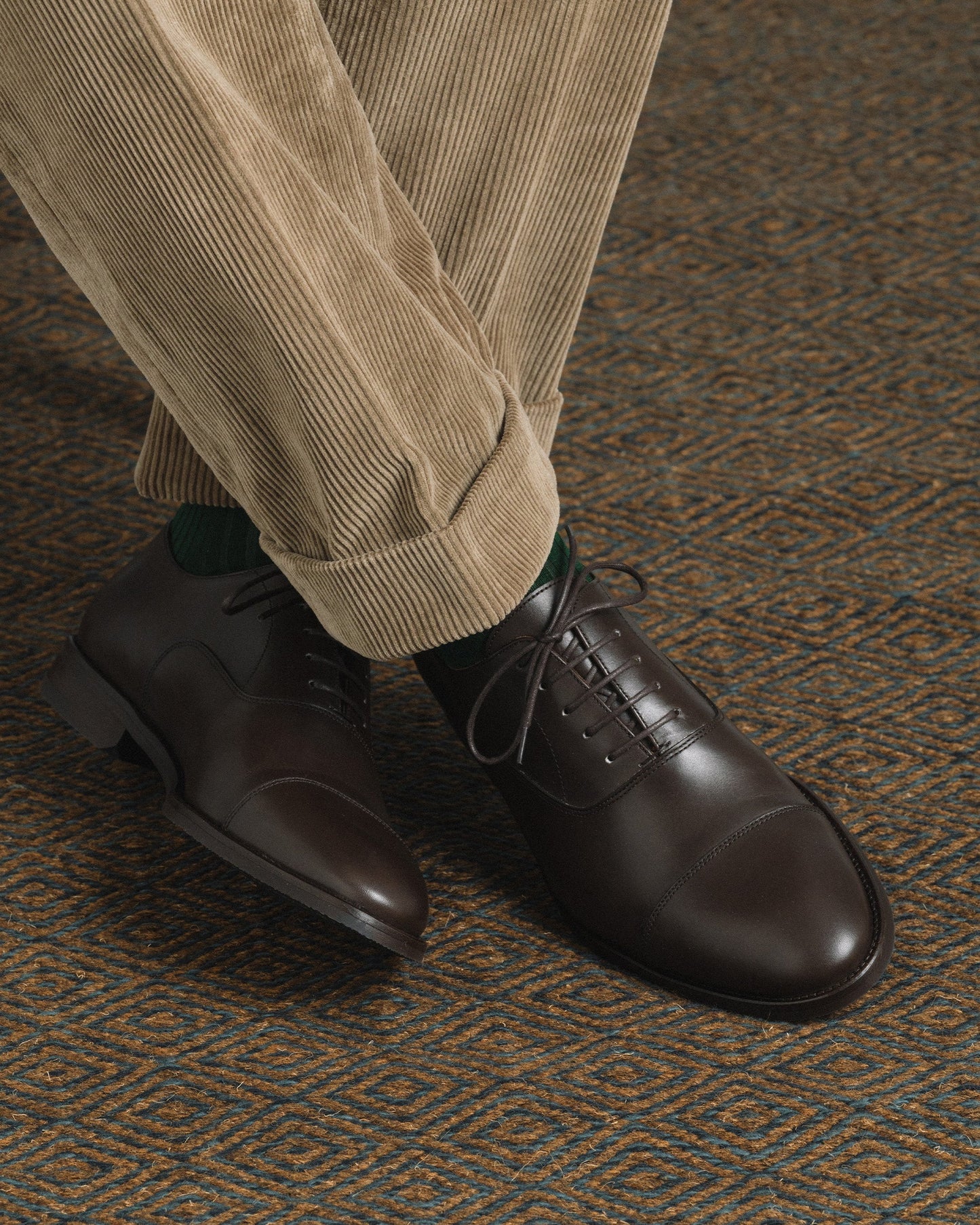 Vinterviken II in dark brown calf, a classic oxford shoe with a chiseled cap toe and clean lines, styled with beige corduroy trousers and green socks on a patterned carpet.